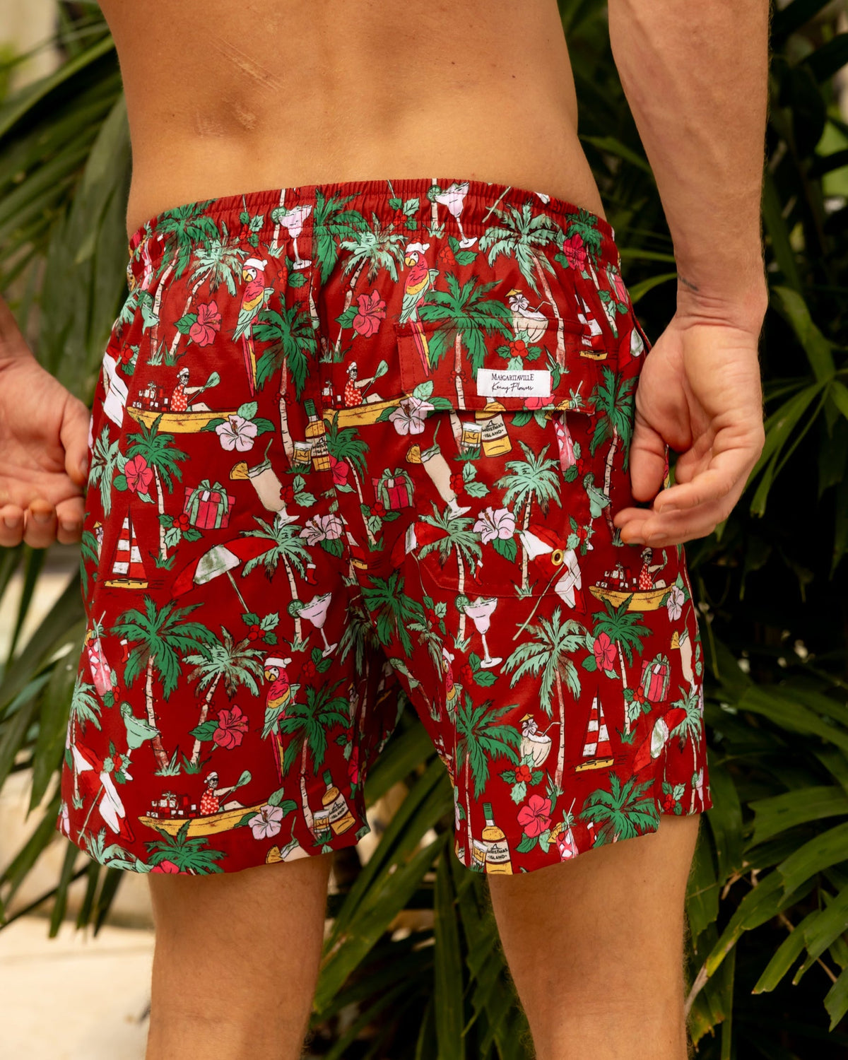Person wearing red tropical patterned holiday swim trunks with plants in the background